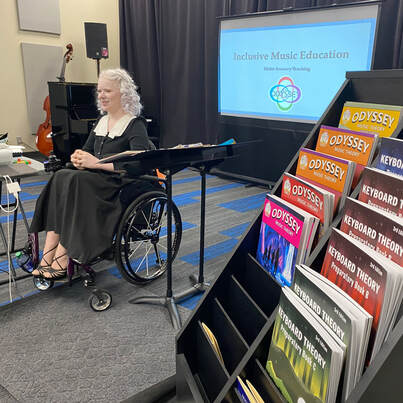 Tamara sits in front of a keynote presentation in her wheelchair. Next to her are a display of books that say Odyssey Music Theory with bright colourful covers.
